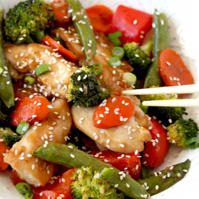 Pictured here a white bowl filled with Asian Chicken and vegetables Stir Fry garnished with sesame seeds and sliced green onions.