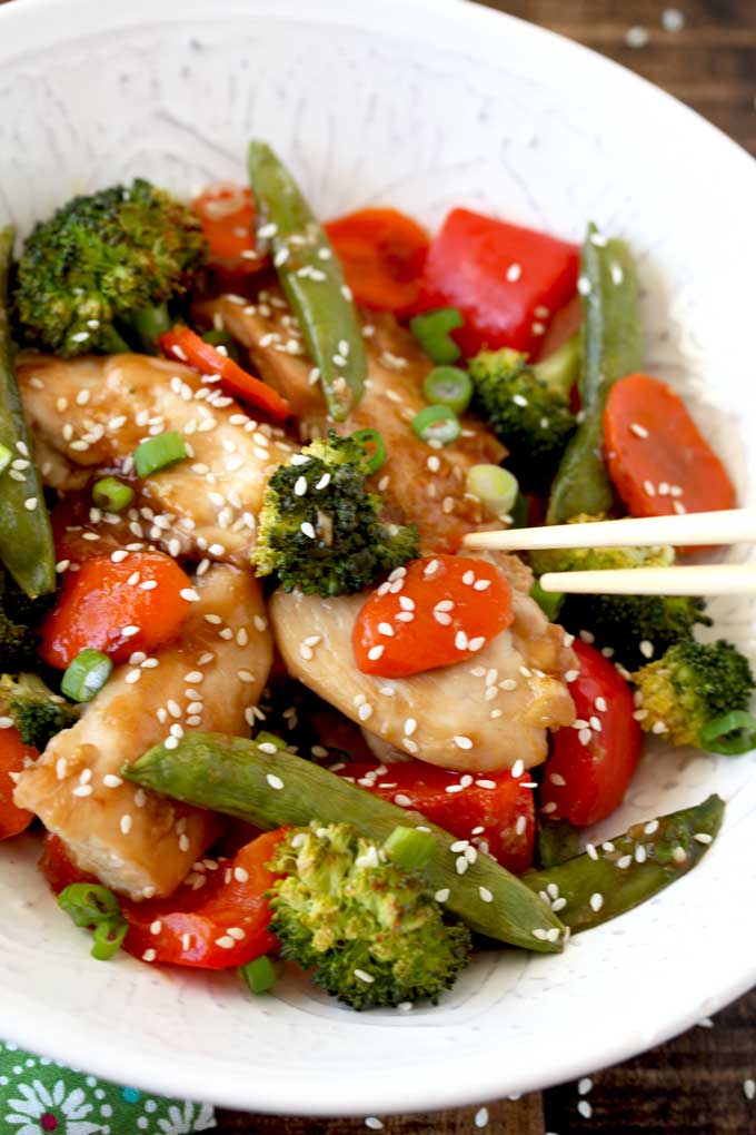 Pictured here a white bowl filled with Asian Chicken and vegetables Stir Fry garnished with sesame seeds and sliced green onions.