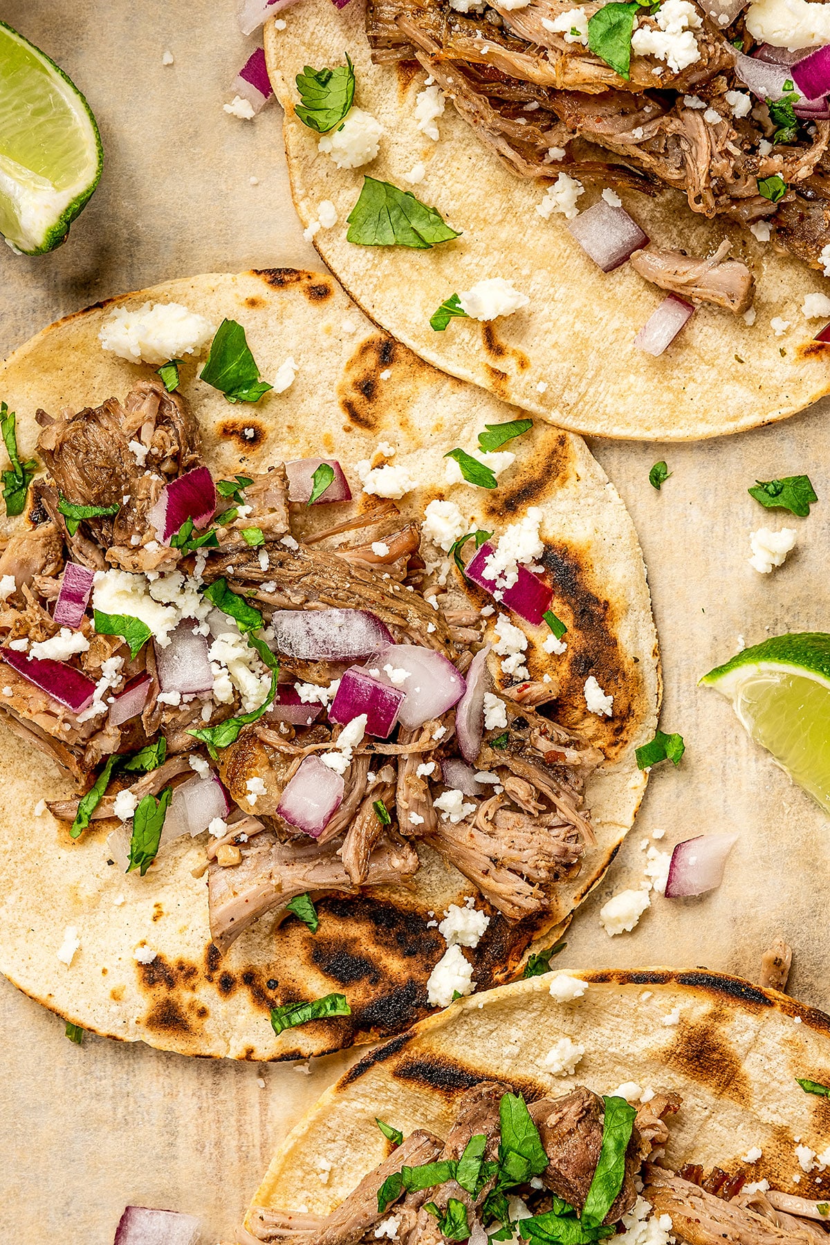 Pictured here a white serving dish filled with shredded slow cooker carnitas, flour tortillas, lime slices and garnished with chopped cilantro.