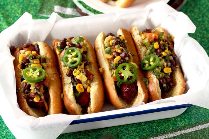 A tray filled with four Southwest Bacon Wrapped Hot Dogs garnished with cilantro and jalapeno slices set on a green tablecloth