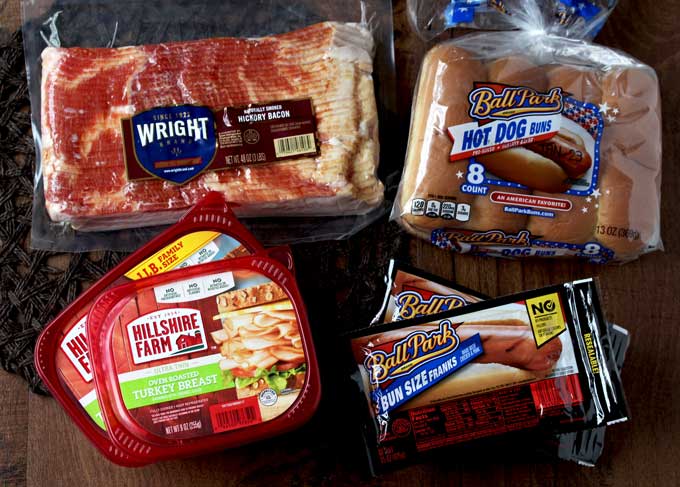 In this photo a package of bacon, a package of Ballpark Hot Dogs and hot dog buns and a package of Hillshire Farms Oven Roasted Turkey