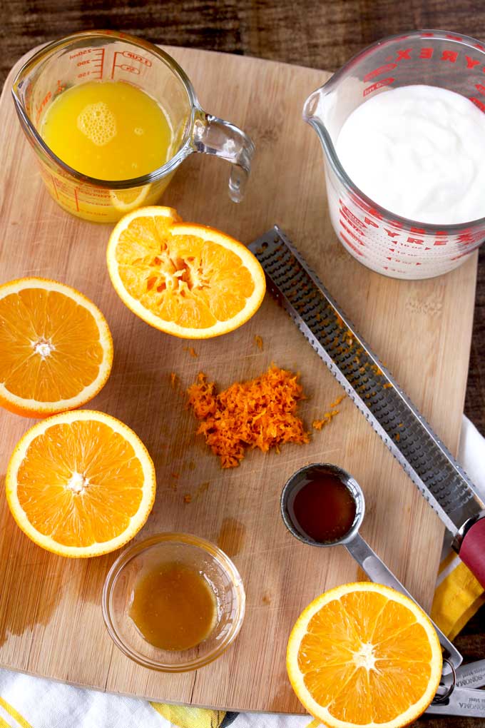View of ingredients to make Orange Honey Lassi on a cutting board. Oranges, orange zest, orange juice, honey and yogurt.