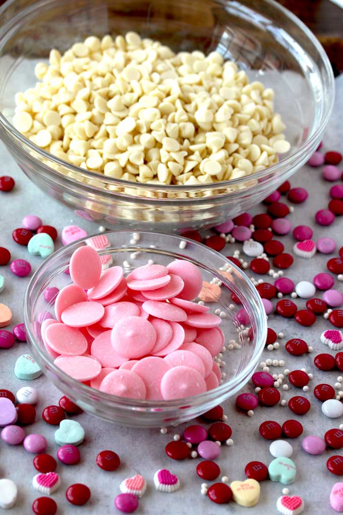 3 ingredients to make the Valentines bark on a sheet pan. A bowl with white chocolate chips, a bowl with pink candy melts and assorted valentine's candy.