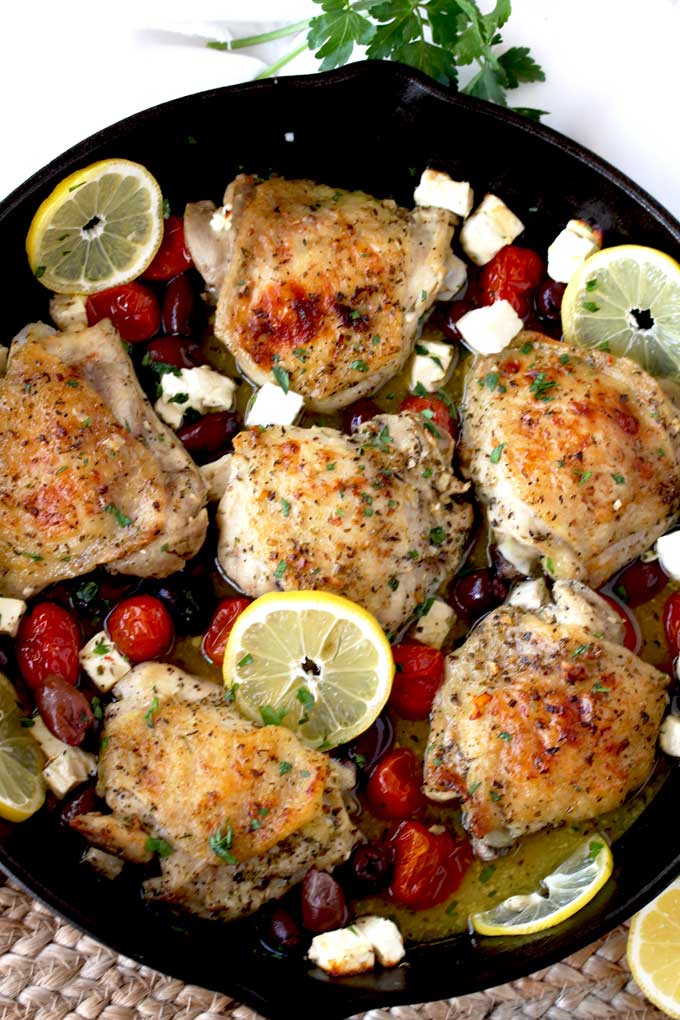 Top view of a cast iron skillet filled with Greek Lemon Oven Roasted Chicken Thighs with tomatoes, Kalamatas, Feta cheese and lemon slices.