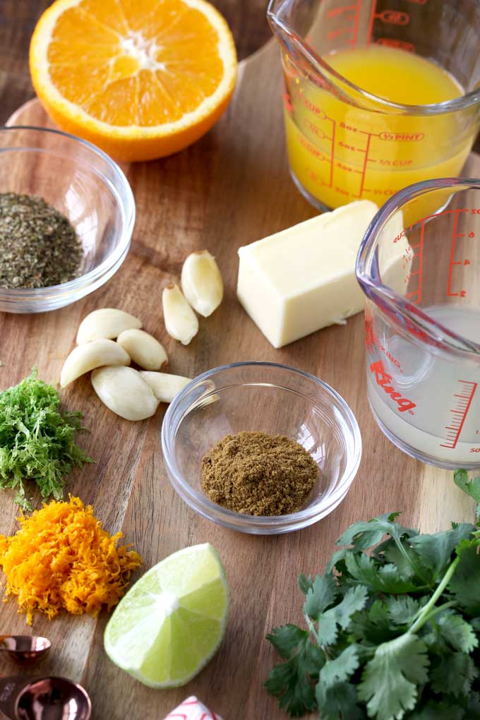 Ingredients to make Mojo Chicken on a wooden board. Lime juice, orange juice, orange and lime zests.=, cilantro, garlic, cumin, oregano and butter.