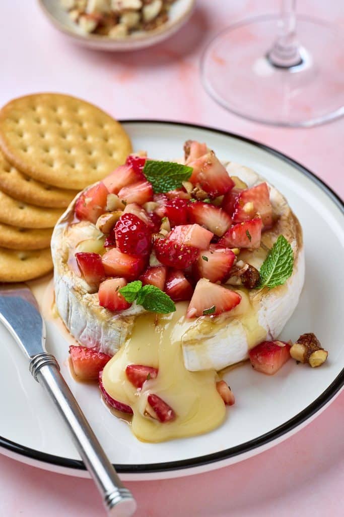 a wheel of oozing brie cheese topped with strawberries and mint on a red baking dish.