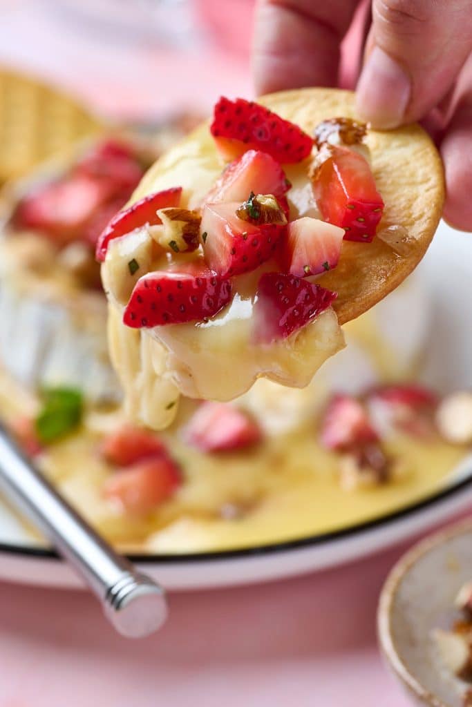 a close up view of a cracker holding melted brie cheese and a few chopped strawberries and candied pecans