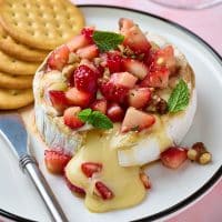 A wheel of warm brie topped with strawberries and mint on white plate.