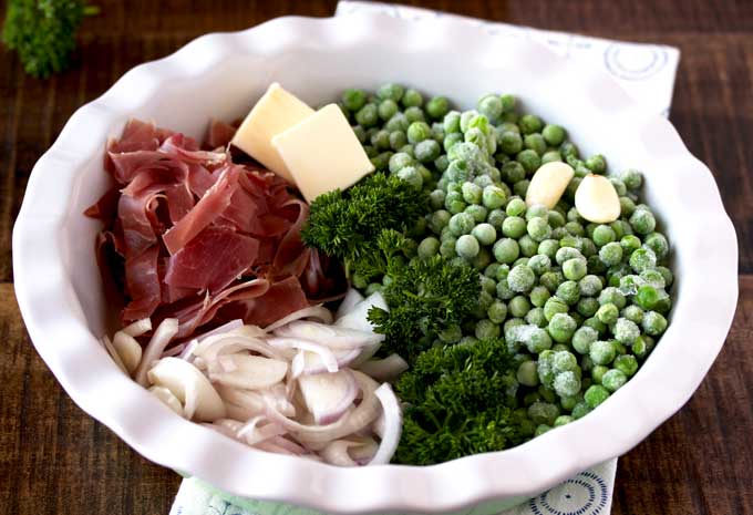 A white plate containing the ingredients to make sauteed peas with prosciutto and shallots. Butter, frozen peas, prosciutto, shallots and chopped parsley.