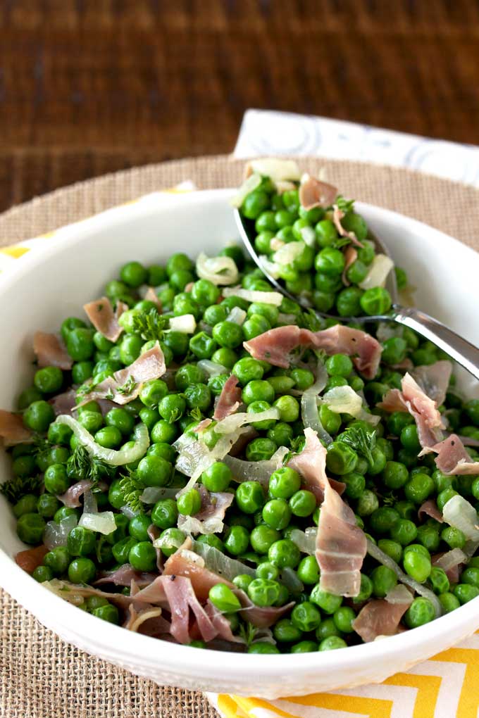 A white bowl filled with sauteed peas with prosciutto and shallots
