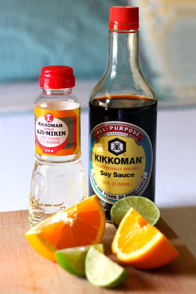 Bottles of cooking sweet rice wine and soy sauce next to fresh lime wedges and fresh orange wedges on a wooden board.