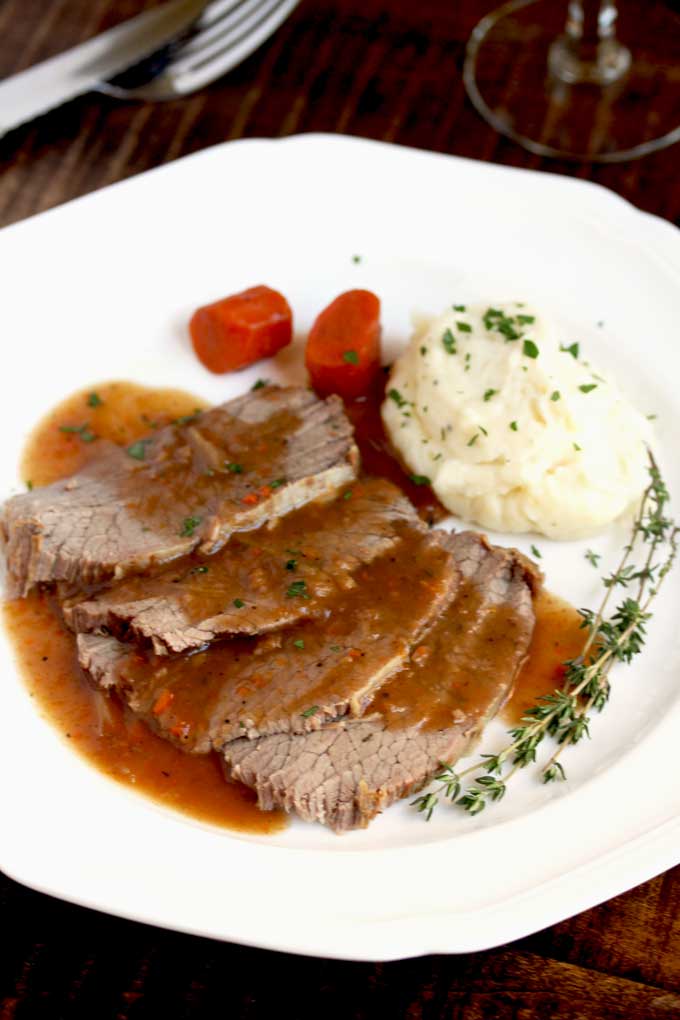 Slices of sauerbraten with mashed potatoes and carrots on a white plate.