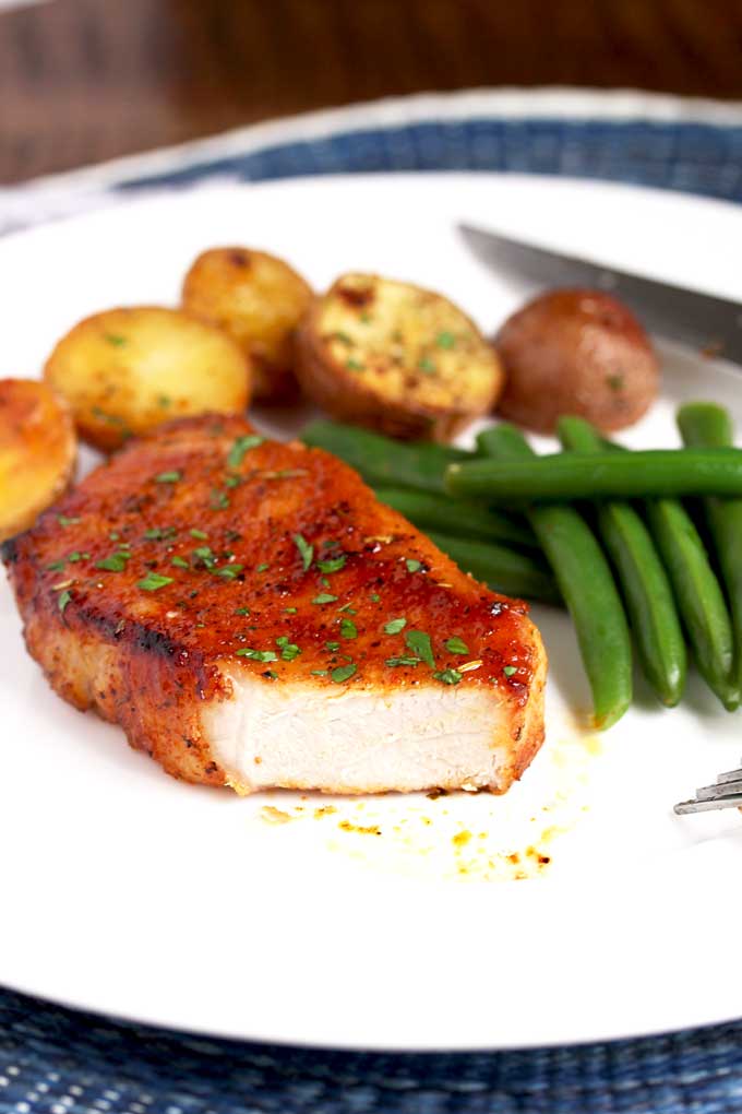 An oven baked pork chop that has been cut on a plate with roasted potatoes and green beans.