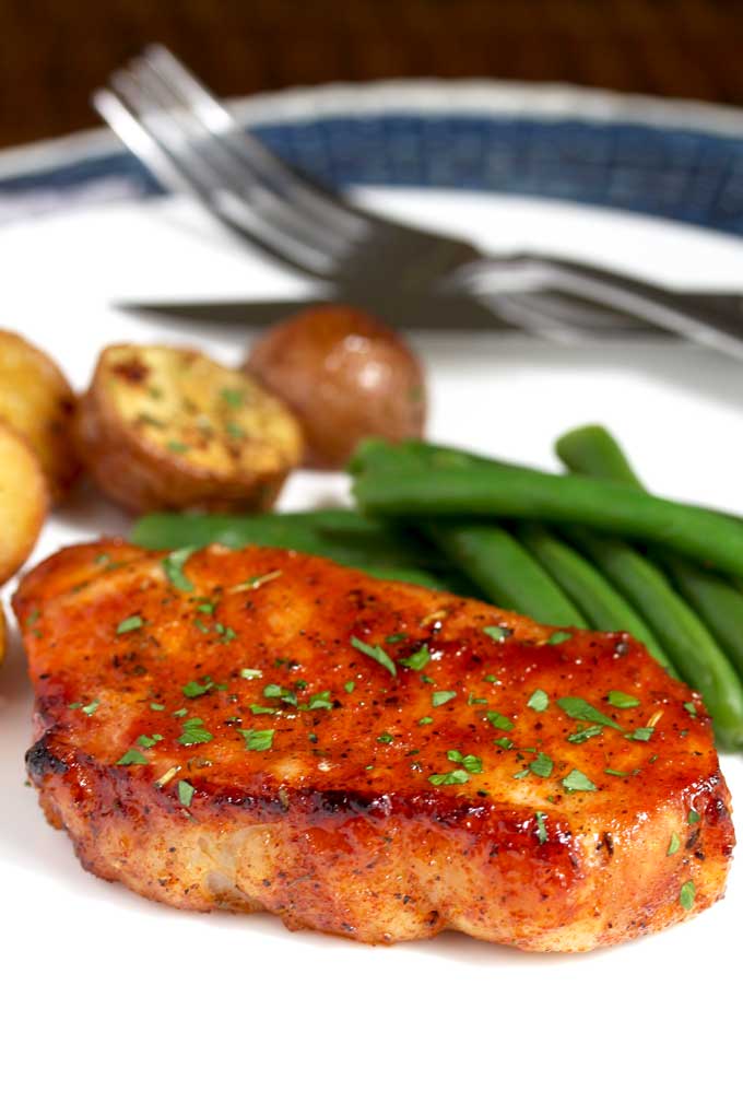 Close up of an oven baked pork chop on a white plate with roasted potatoes and green beans.