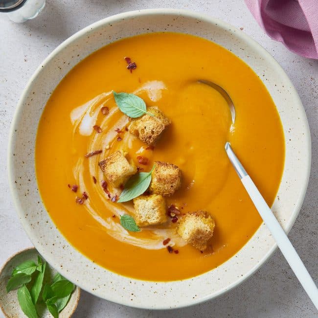 A soup bowl filled with creamy carrot soup and topped with croutons, red pepper flakes and fresh herbs and drizzled with coconut milk.