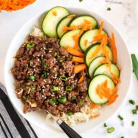 Korean ground beed over rice with cucumbers and carrots served in a white bowl.