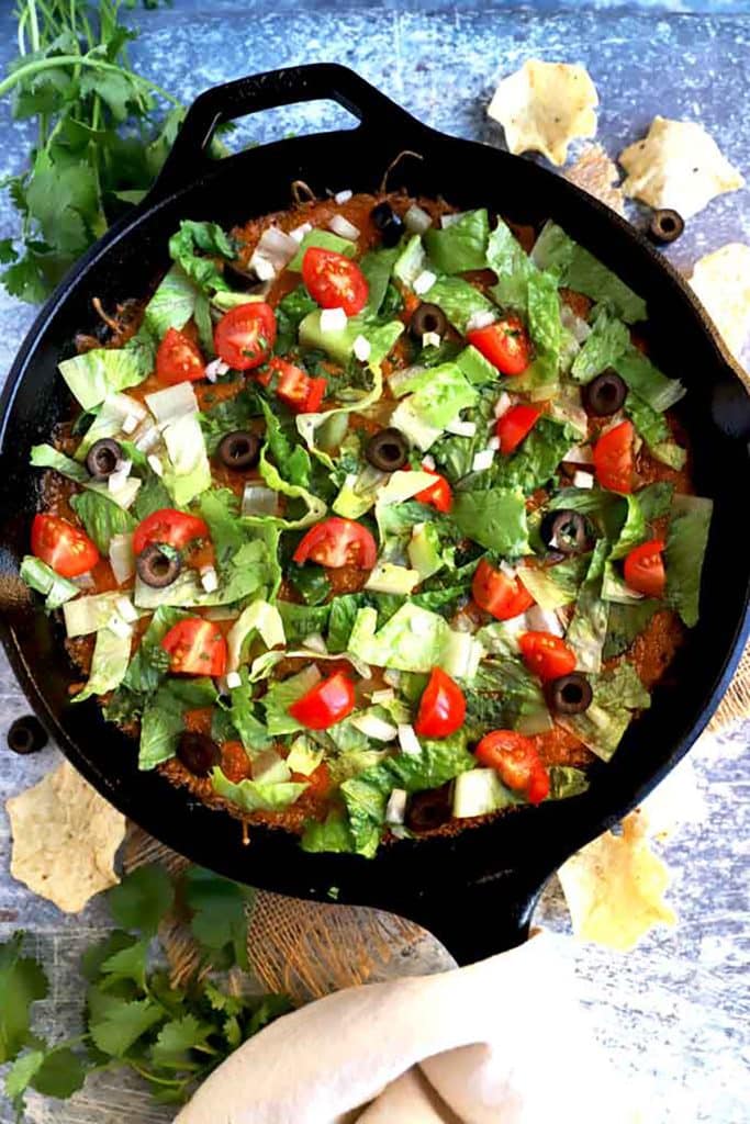 View of a skillet with taco dip topped with lettuce, tomatoes and olives.