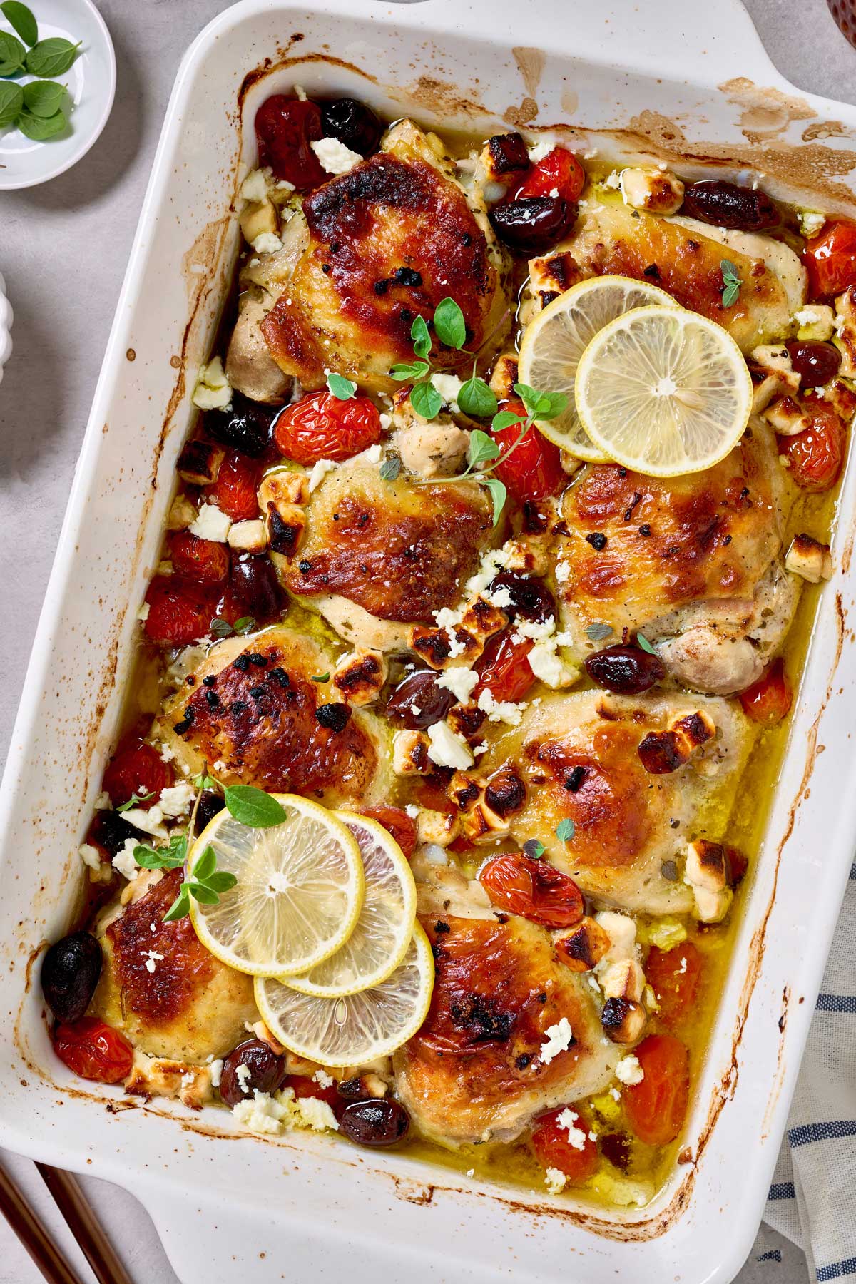 Golden-browned Greek chicken thighs baked in a white dish with blistered cherry tomatoes, kalamata olives, crumbled feta, lemon slices, and fresh oregano.