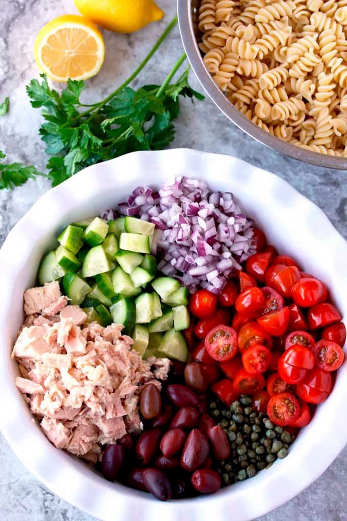 Ingredients to make Tuna Pasta Salad in a bowl. Chopped Onions, Tomatoes, Kalamata olives, Tuna, cucumbers, capers. In a colander cooked pasta.