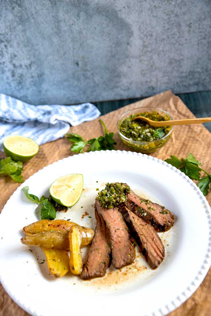 A few sliced of grilled steak topped with chimichurri served with golden potatoes on a white plate.