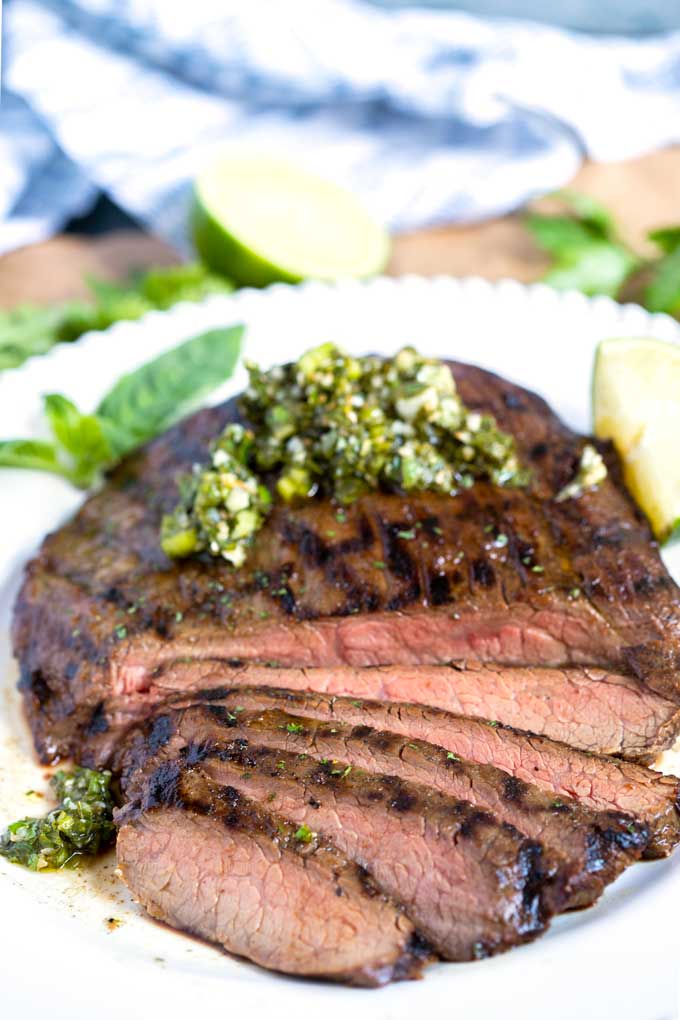 Sliced grilled flank steak topped with herb chimichurri on a white platter.