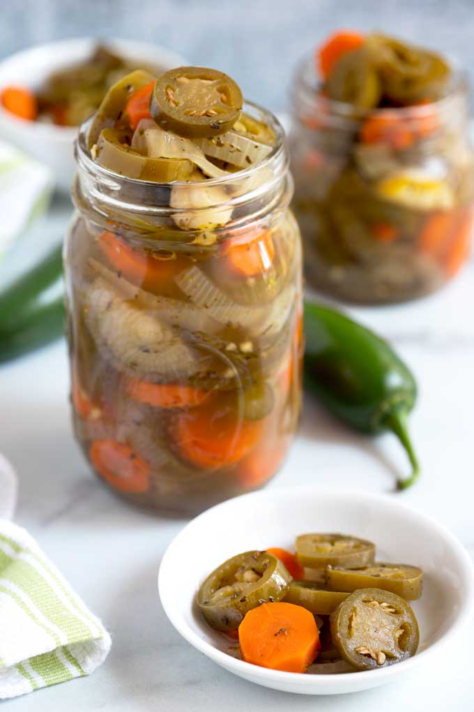 A jar filled to the rim with pickled jalapenos and carrots next to a small plate with pickled jalapenos.