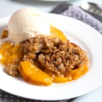 Peach dessert with crumbly topping on a white plate served with vanilla ice cream.