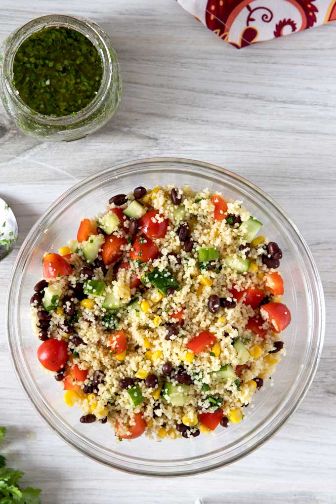 Mixing Couscous Salad in a glass mixing bowl next to a jar of cilantro lime vinaigrette.