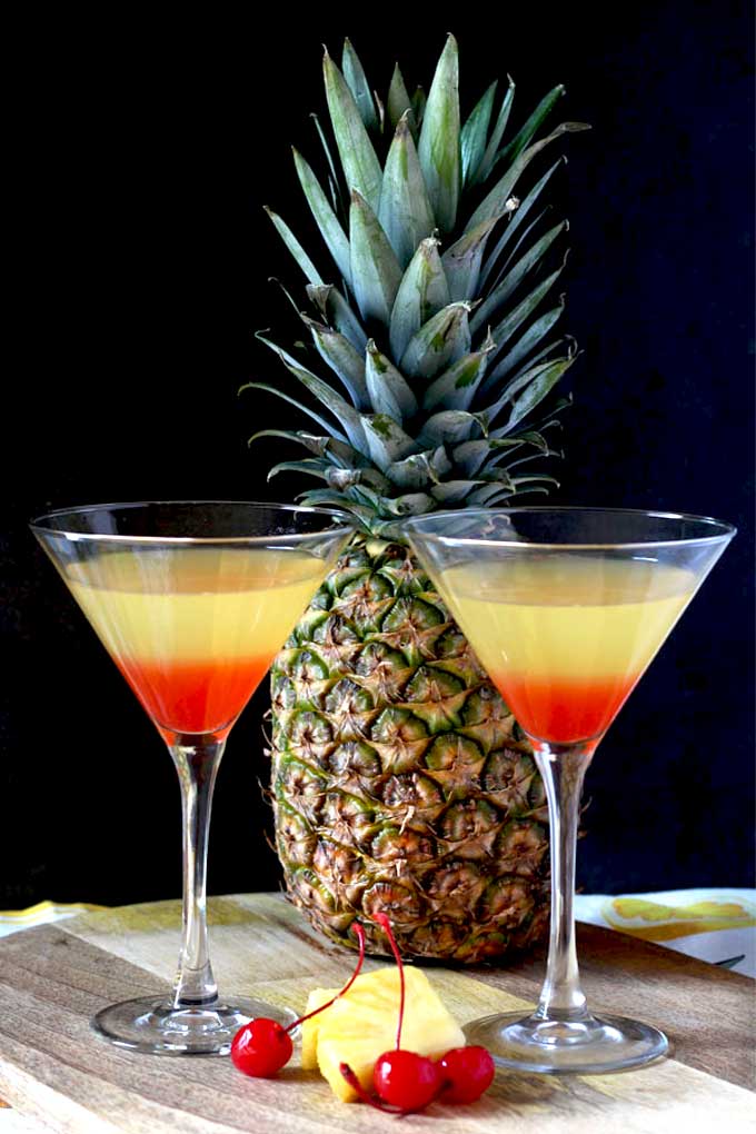 2 martini glasses with tropical Bikini martini, pineapple and maraschino cherries on a wooden board.