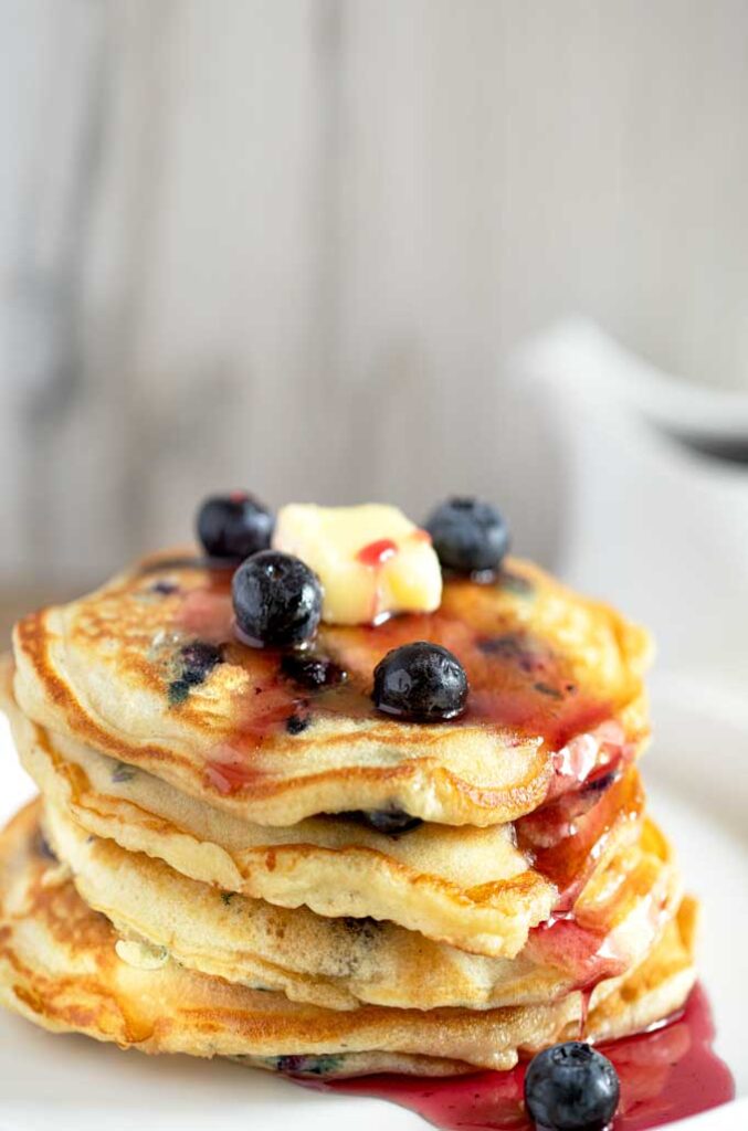 A stack of blueberry pancakes with butter and blueberry syrup on a white plate.