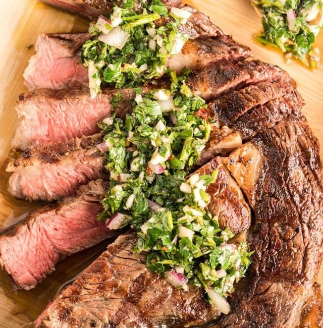 Sliced ribeye steak topped with Chimichurri on a cutting board.