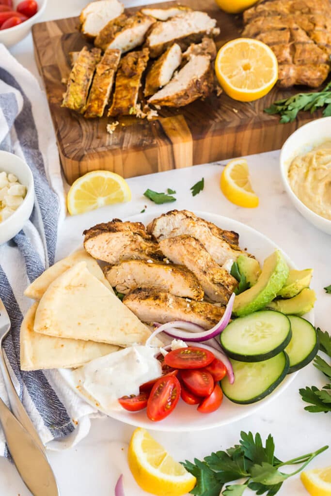 A plate with slices of chicken shawarma, cucumbers, onions, tomatoes, avocado, pita bread and yogurt sauce on a lunch plate.