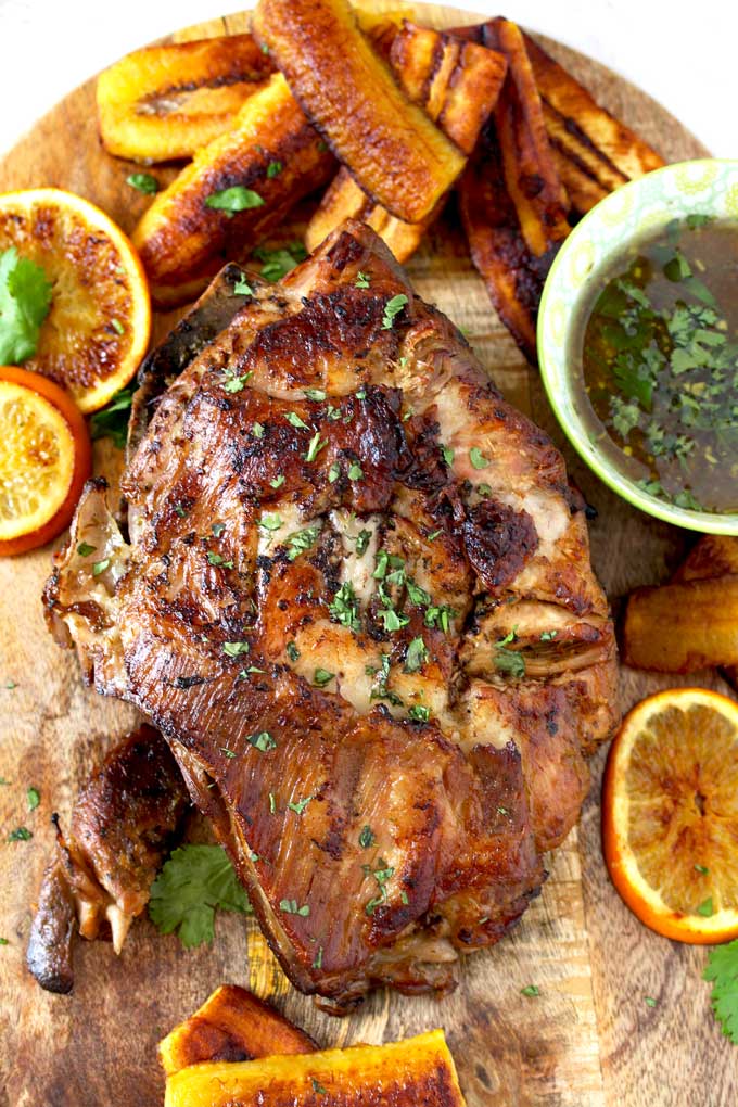 Top view of crispy Cuban Pork Roast on a cutting board surrounded with fried plantains and a bowl of Cuban Mojo criollo, 