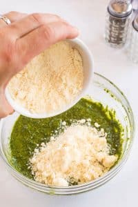 pouring parmesan cheese into a bowl with pesto sauce