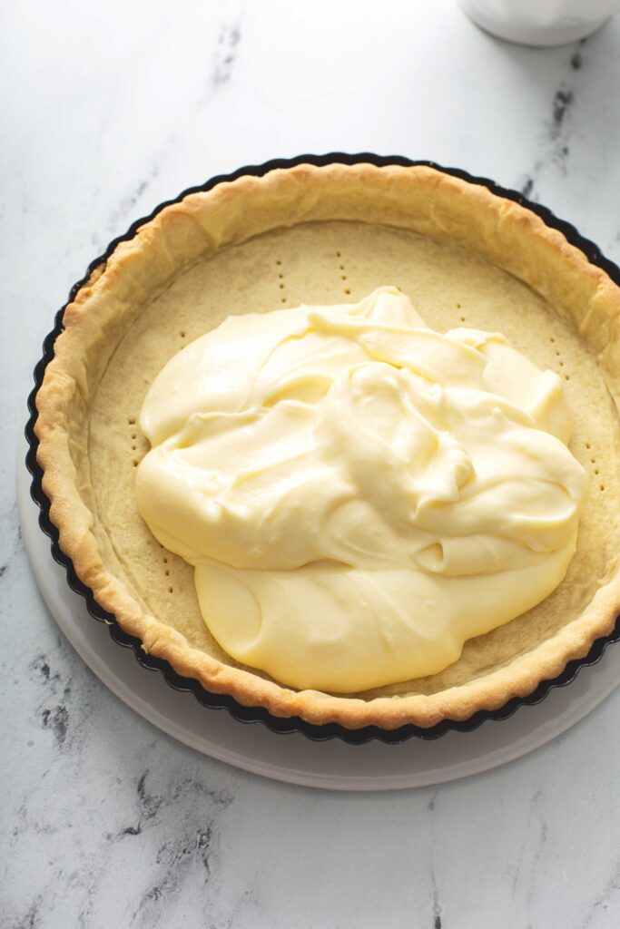 A baked golden brown pastry tart crust getting topped with vanilla pastry cream.