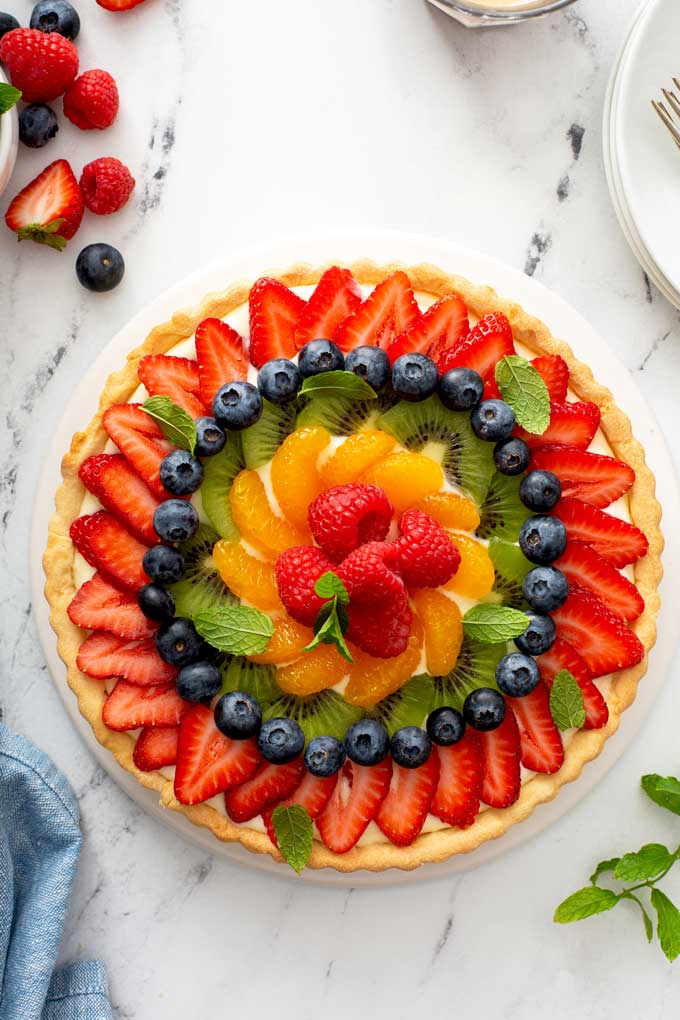 A French Fresh Fruit Tart with strawberries, kiwi, mandarin oranges, raspberries and blueberries on a white platter.