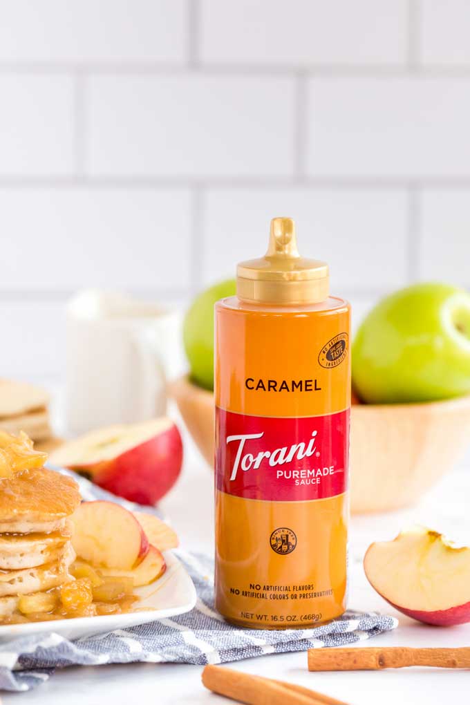 A bottle of Torani Caramel Sauce next to apples, cinnamon and pancakes.