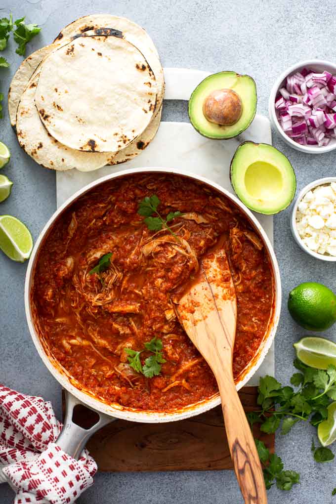 A pot filled with Chicken Tinga surrounded by corn tortillas, limes, avocado, cilantro and onions.