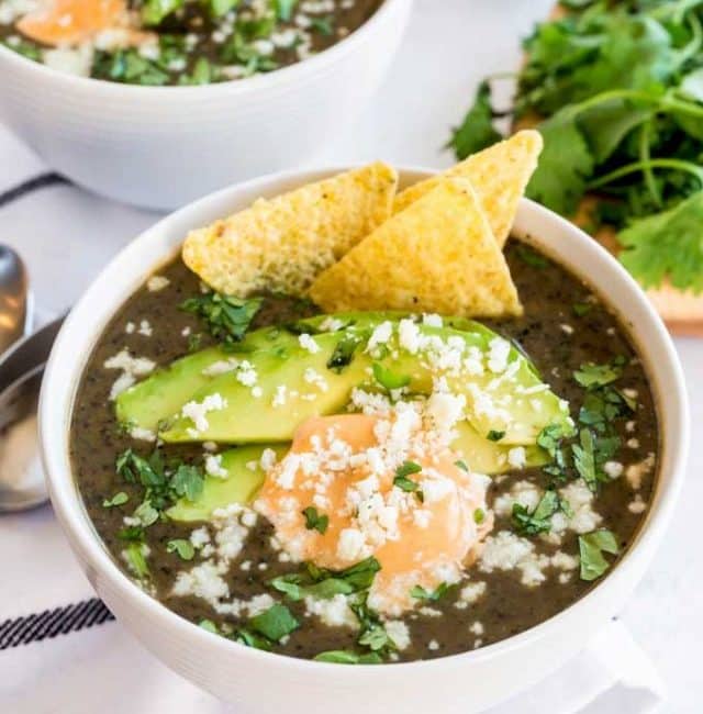 Black bean soup topped with chipotle crema, avocados, cheese, cilantro and tortilla chips in a white bowl.