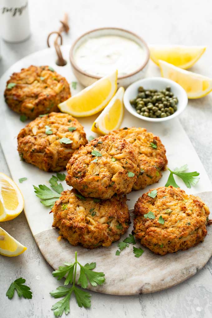 Crab cakes on a white marbled board with a bowl of lemon aioli and capers.