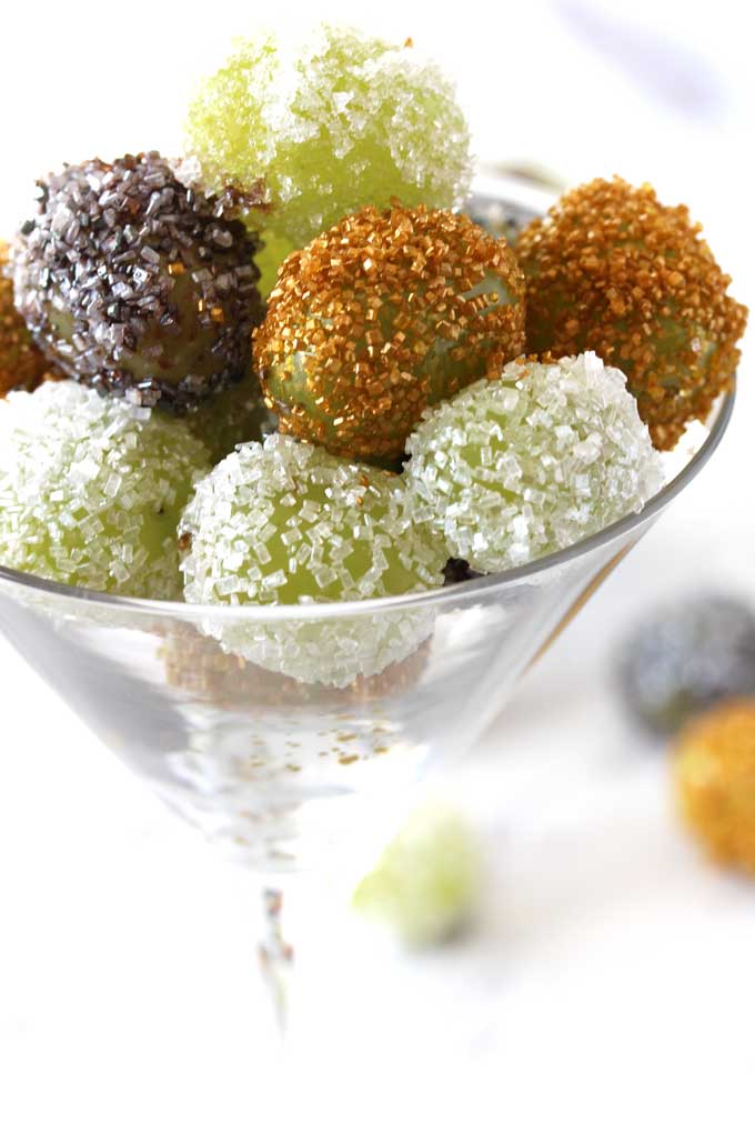 Close up view of a Martini glass filled with sugared grapes in gold, white and silver next to a small square glass bowl filled also with candied grapes