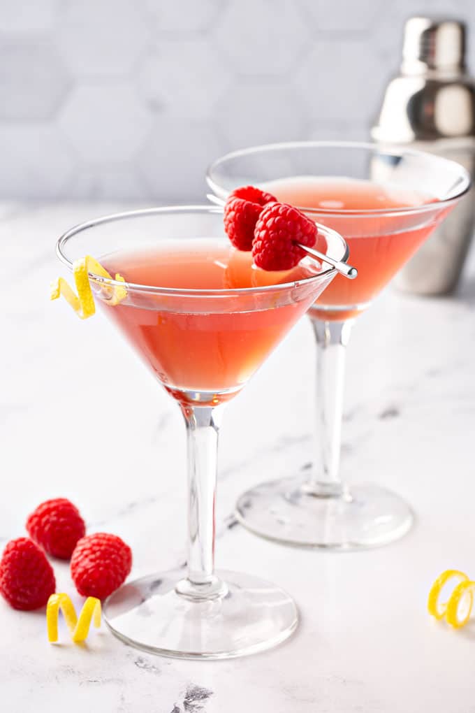 Two martini glasses with a martini garnished with fresh raspberries and a lemon twist.