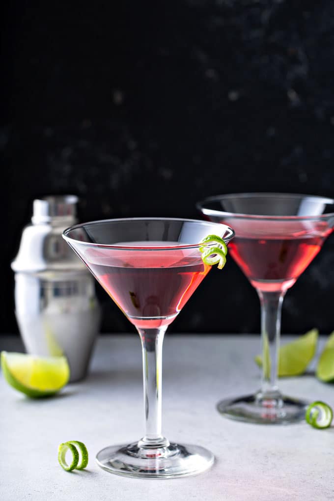 A couple of cosmopolitan cocktails on a white surface next to a cocktail shaker