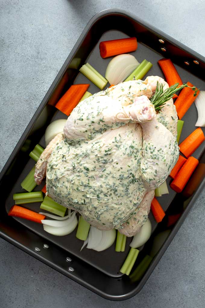 A whole chicken slathered with herb butter in a roasting pan over vegetables.