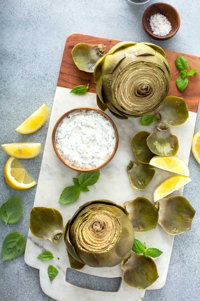 two artichokes with dipping sauce in a small bowl over a marble board