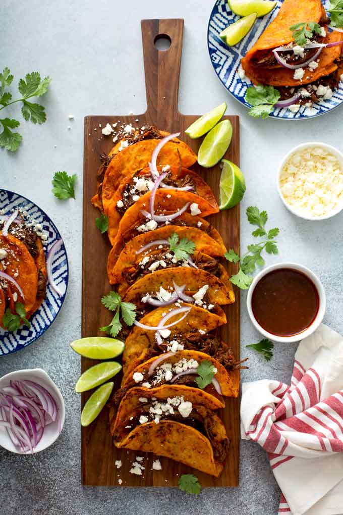 Several Birria Tacos on a wooden board with consommé  surrounded by plates filled with tacos