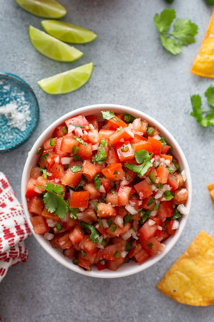 Top view of a white bowl with Pico de Gallo next to slices of lime and chips
