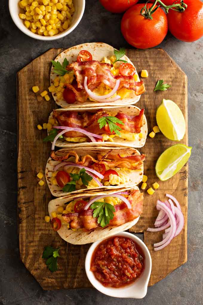 Multiple breakfast tacos with eggs and bacon on a wooden board.