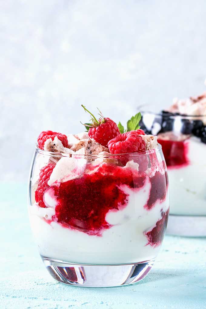 Glass with whipped cream, raspberries and meringue on a light blue surface.