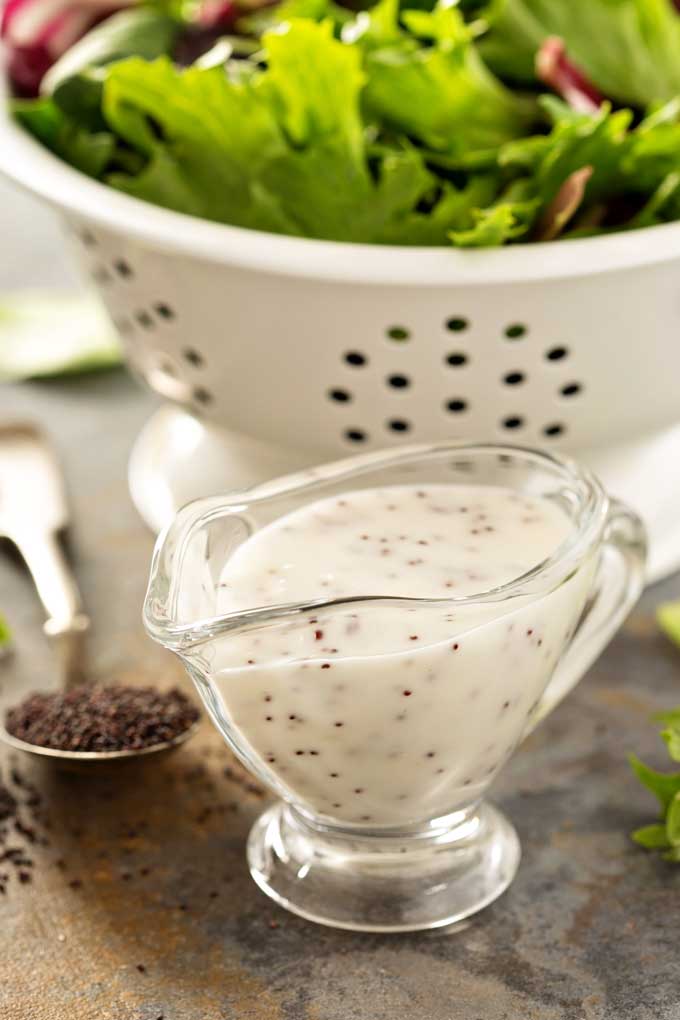 Lemon Poppy seed Dressing in a small glass pitcher next to a colander filled with salad greens